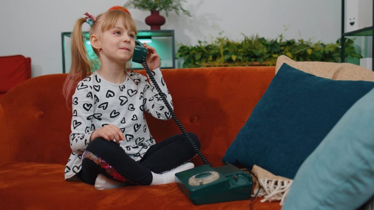 Smiling young little teen children girl making fun conversation call on landline retro telephone with friends, sitting on couch at home. Happy excited toddler child kid enjoying talking communication